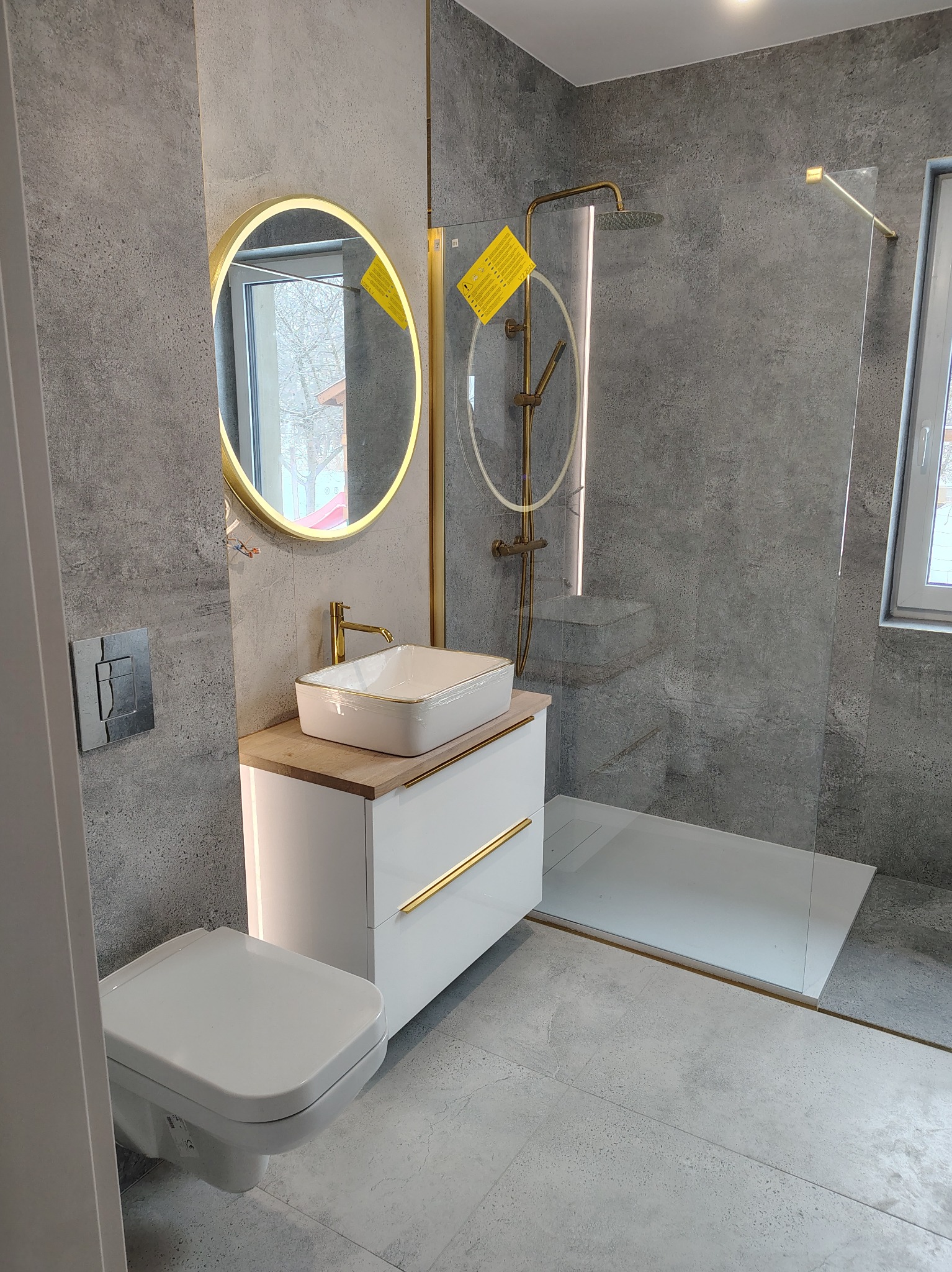 Nowoczesna łazienka z szarą płytką, okrągłym lustrem z podświetleniem LED, białą szafką z umywalką i złotymi akcentami oraz kabiną prysznicową typu walk-in.