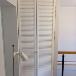 Yauheni Smirnou - Biała szafa wnękowa z żaluzjowymi drzwiami, wkomponowana w przestrzeń przy schodach, oświetlona lampą stojącą.