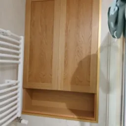 Drewniana szafka łazienkowa z dwoma drzwiczkami i otwartą półką poniżej, zamontowana nad spłuczką, obok grzejnika drabinkowego.