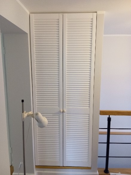 Biała szafa wnękowa z żaluzjowymi drzwiami, wkomponowana w przestrzeń przy schodach, oświetlona lampą stojącą.