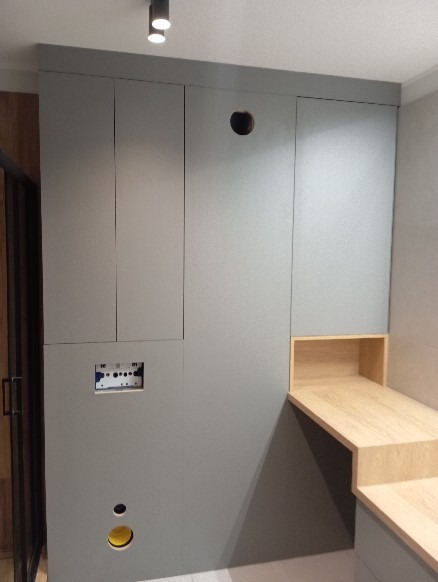 Szara zabudowa meblowa na wymiar z drewnianym blatem, widoczne otwory na instalacje i oświetlenie punktowe.