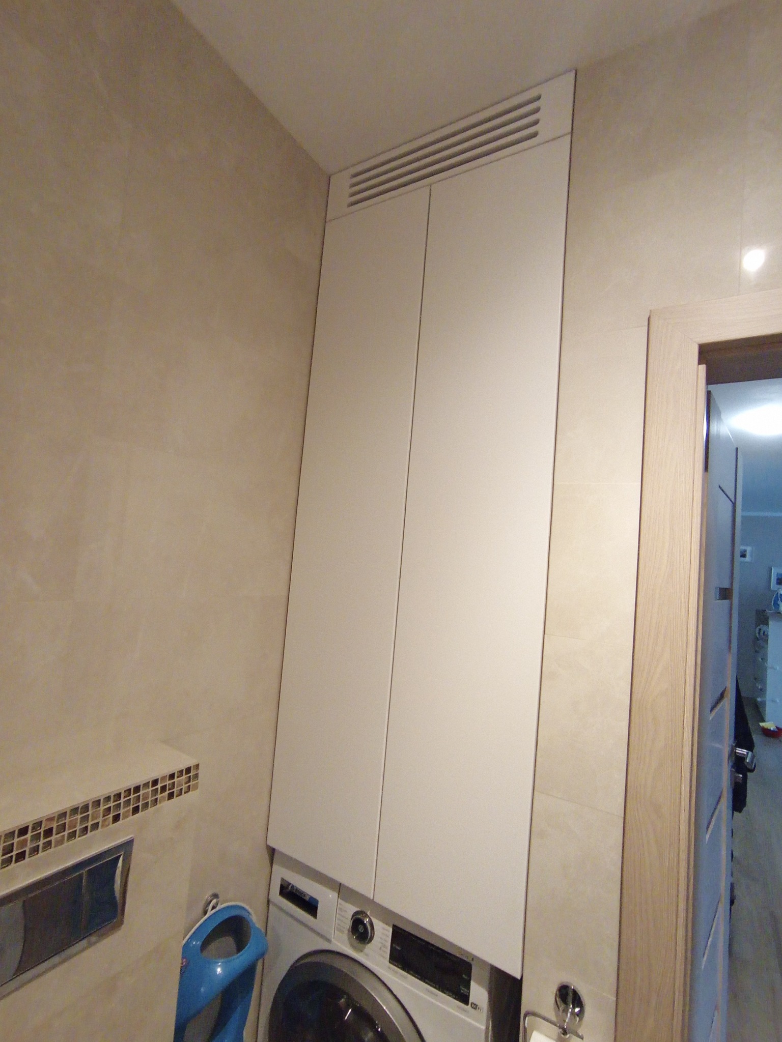 Biała szafa wnękowa z wentylacją u góry, zabudowana nad pralką w łazience z beżowymi płytkami.