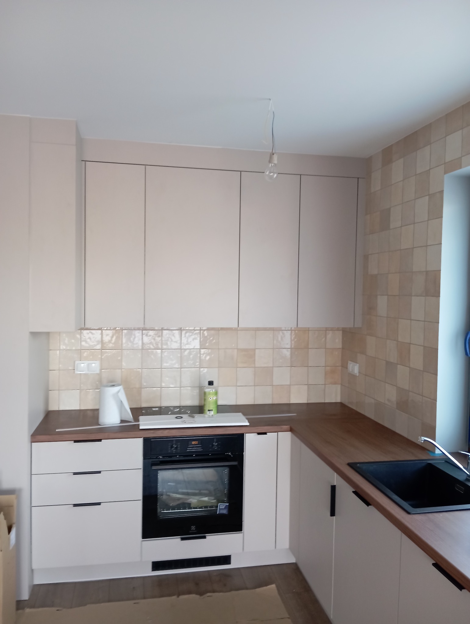 Jasna kuchnia z drewnianym blatem, beżowymi szafkami i piekarnikiem Electrolux, płytki w odcieniach beżu na ścianie, czarny zlew i bateria.