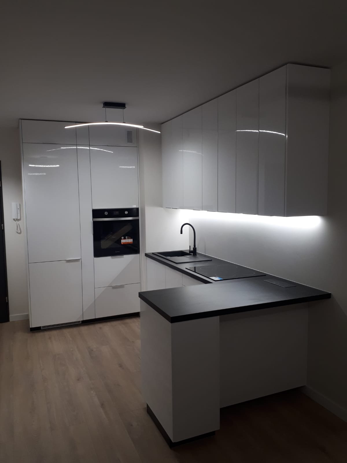 Nowoczesna, minimalistyczna kuchnia z białymi, lakierowanymi frontami, czarnym blatem i podświetleniem LED pod szafkami wiszącymi oraz lampą wiszącą w kształcie łuku.