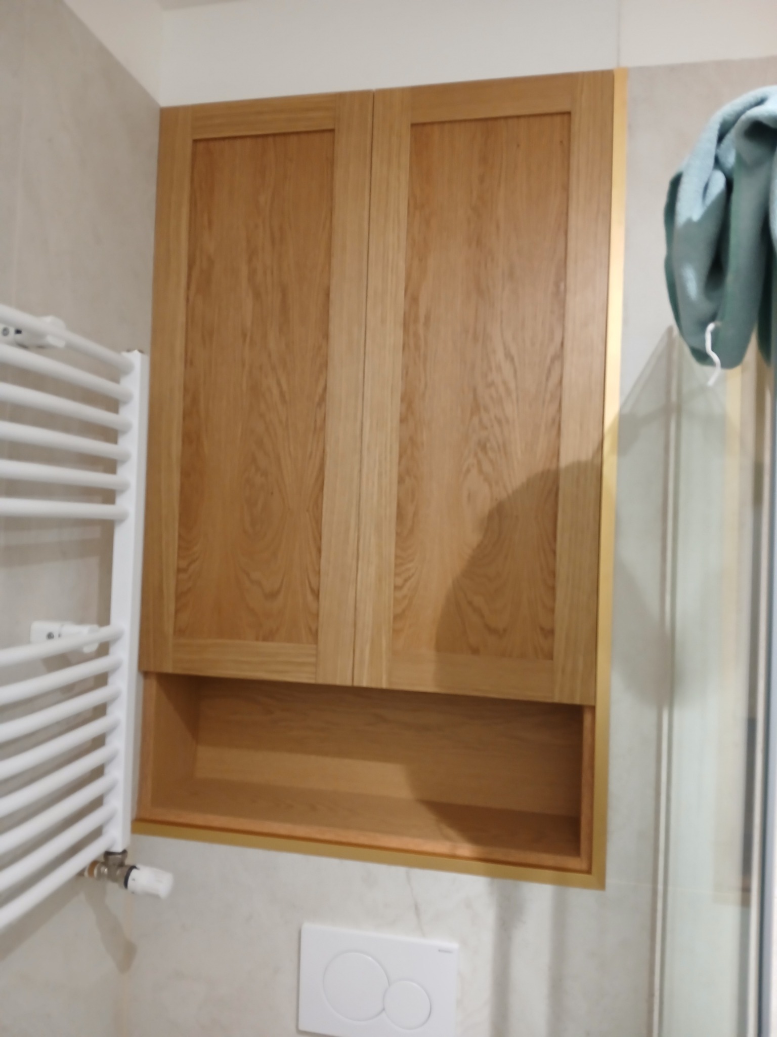 Drewniana szafka łazienkowa z dwoma drzwiczkami i otwartą półką poniżej, zamontowana nad spłuczką, obok grzejnika drabinkowego.