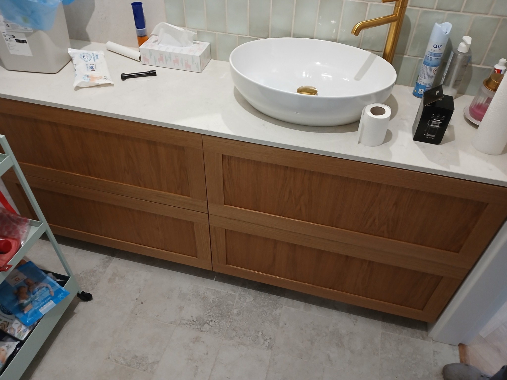 Łazienkowa szafka z drewnianymi frontami pod umywalką nablatową, widoczne akcesoria łazienkowe i fragment wózka z akcesoriami dla dzieci.