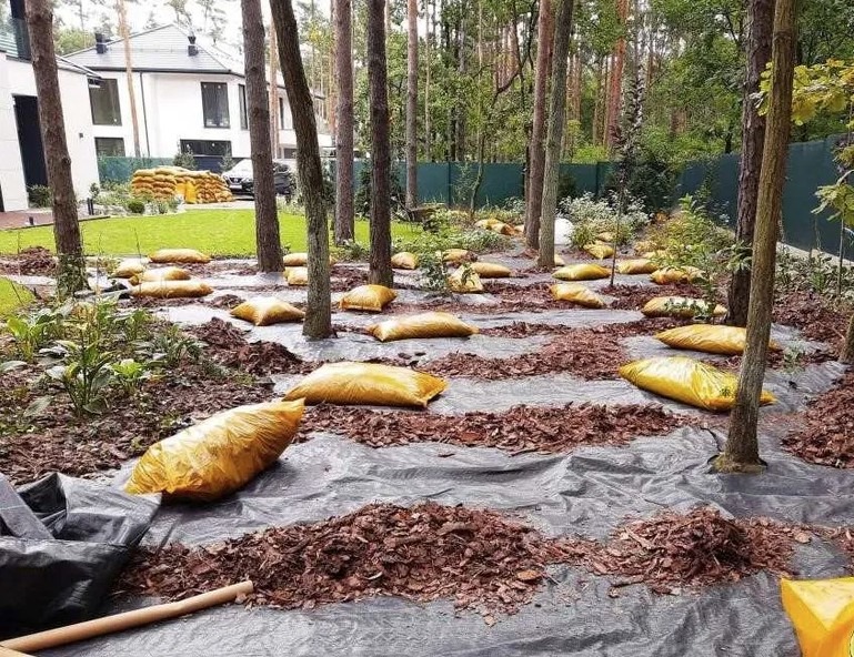 Przygotowanie terenu pod trawnik: rozłożona agrowłóknina, worki z ziemią i kora rozsypana między drzewami, w tle dom jednorodzinny i sterta worków z ziemią.