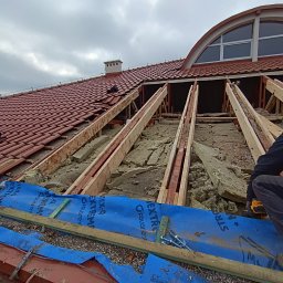 Wojciech Dampc - Rozpoczęte prace przy wymianie pokrycia dachowego z widoczną więźbą, ociepleniem i membraną dachową, fragment czerwonej dachówki i okno w kształcie półkola na tle pochmurnego nieba.