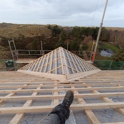 Wojciech Dampc - Widok z góry na konstrukcję drewnianą dachu w trakcie budowy, z widoczną częścią nogi w roboczym bucie, w tle krajobraz zalesionych wydm i morza.