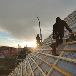 Wojciech Dampc - Dwóch dekarzy pracuje na drewnianej konstrukcji dachu w promieniach zachodzącego słońca, widoczny dźwig i zabudowa jednorodzinna w tle.