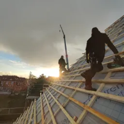 Dwóch dekarzy pracuje na drewnianej konstrukcji dachu w promieniach zachodzącego słońca, widoczny dźwig i zabudowa jednorodzinna w tle.