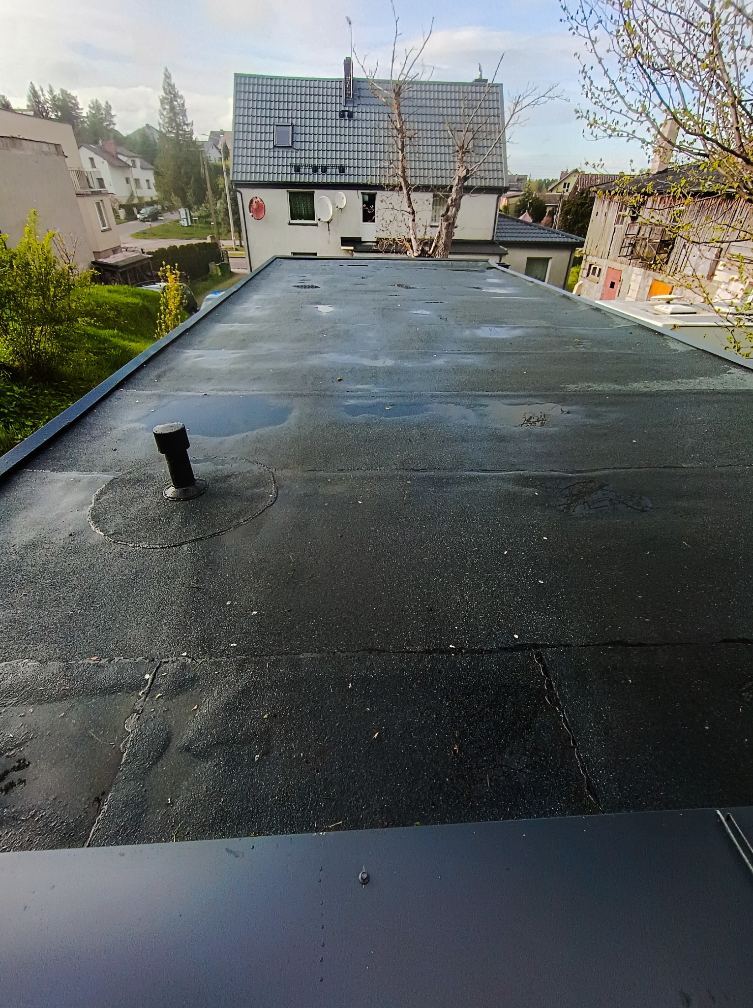 Czarny, płaski dach pokryty papą z widocznym kominkiem wentylacyjnym i kałużami po deszczu, w tle zabudowa jednorodzinna.