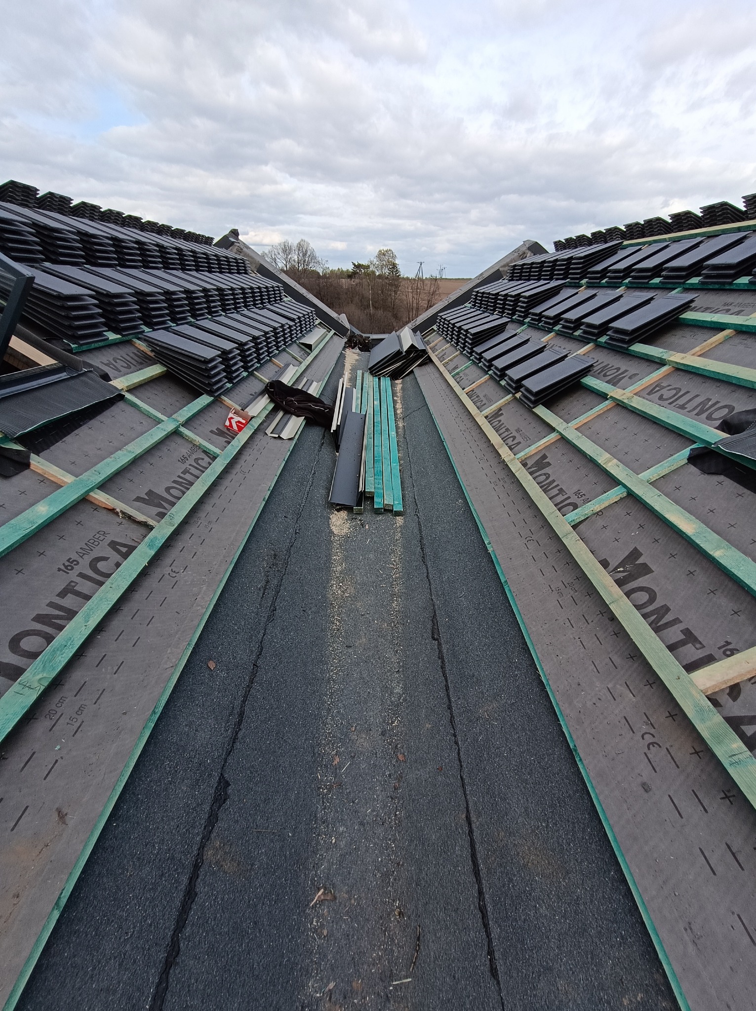 Układanie czarnej dachówki na konstrukcji dachu z widoczną membraną i łatami, widok z góry na rozdzieloną połać dachową, fragmenty desek i akcesoriów dekarskich na dachu.