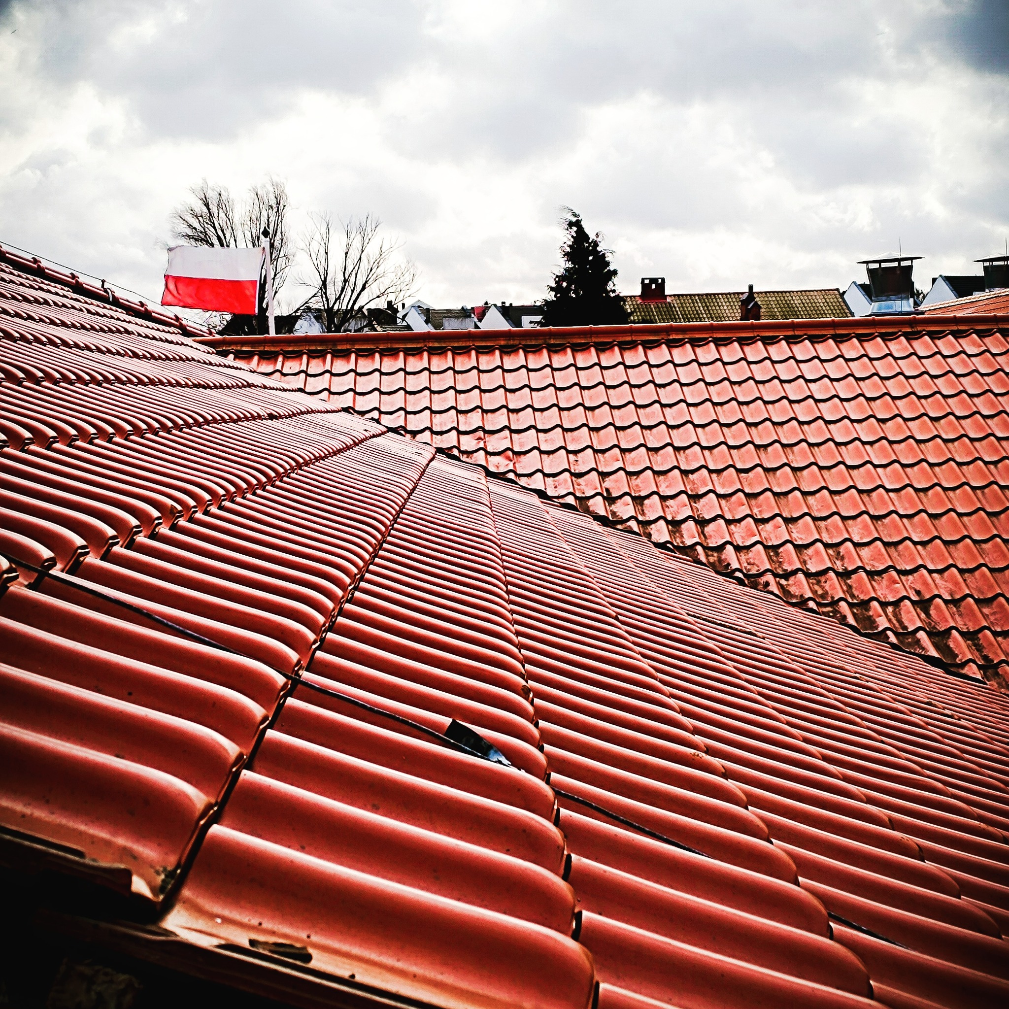 Czerwona dachówka ceramiczna na dachu budynku, z widoczną flagą Polski w tle i pochmurnym niebem.
