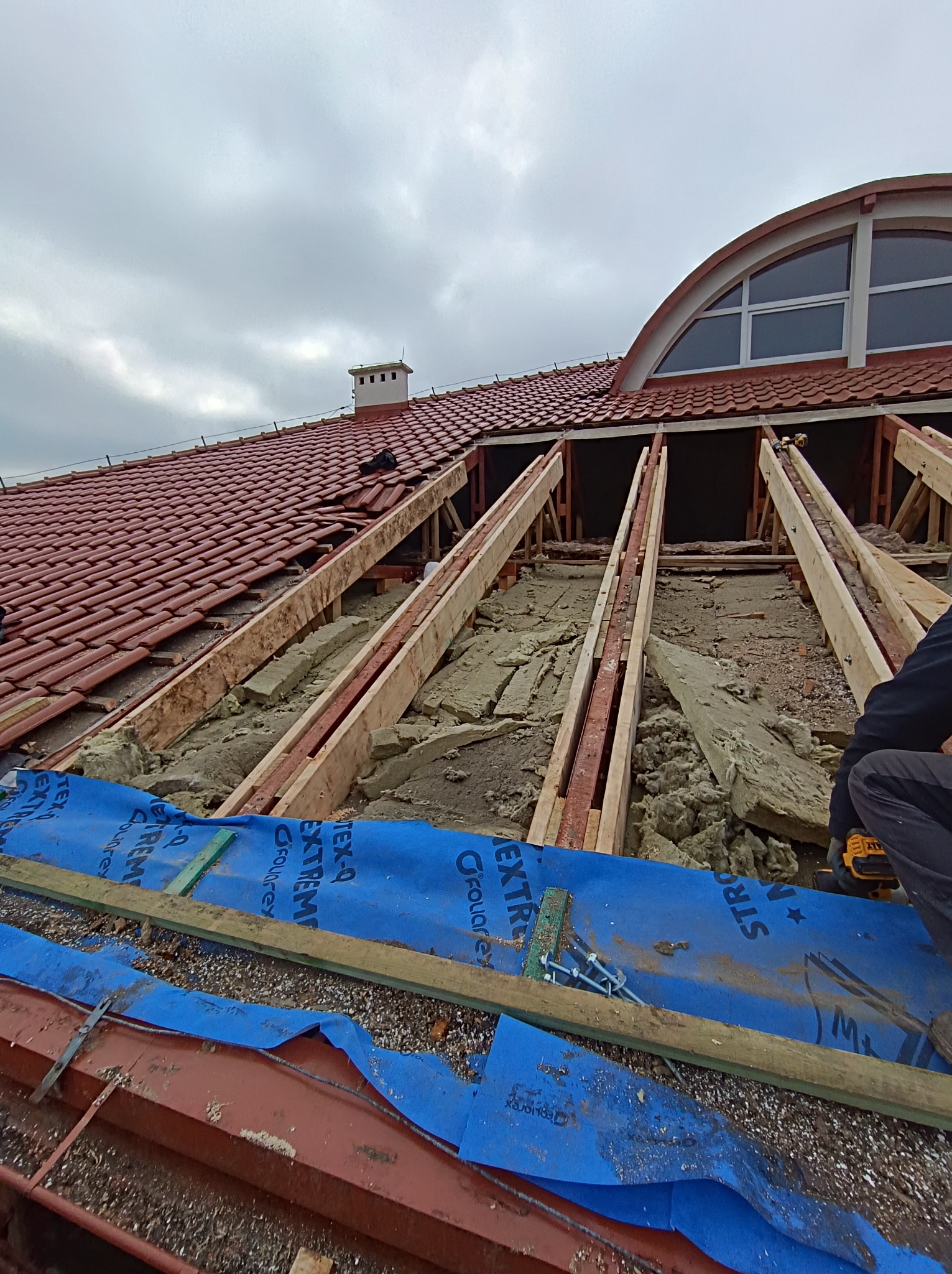 Rozpoczęte prace przy wymianie pokrycia dachowego z widoczną więźbą, ociepleniem i membraną dachową, fragment czerwonej dachówki i okno w kształcie półkola na tle pochmurnego nieba.