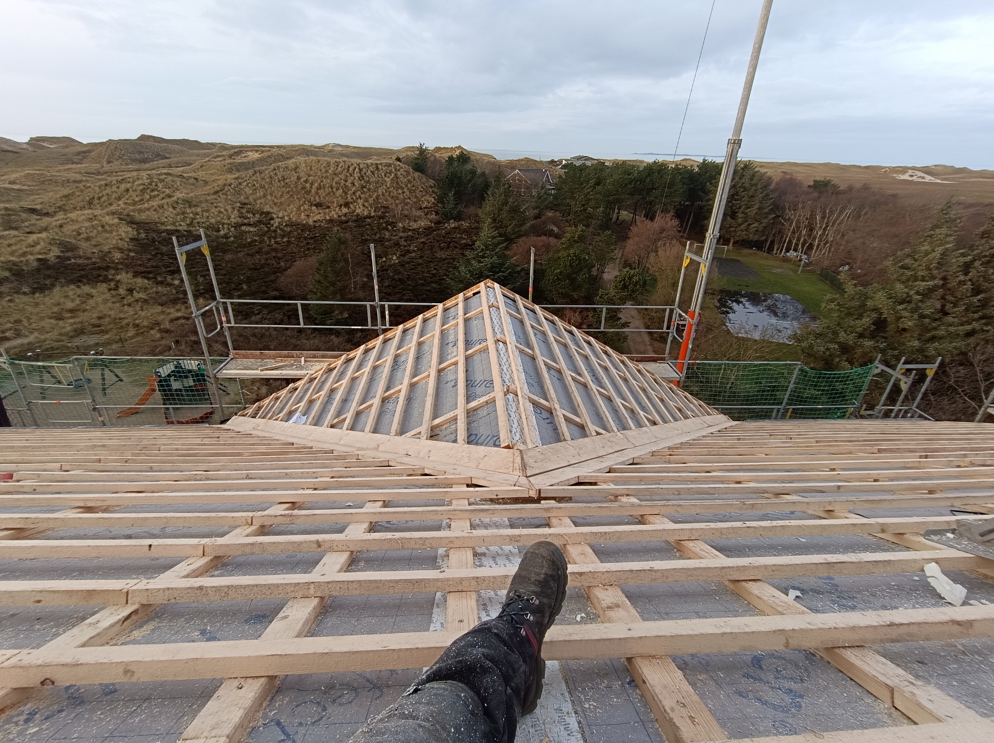 Widok z góry na konstrukcję drewnianą dachu w trakcie budowy, z widoczną częścią nogi w roboczym bucie, w tle krajobraz zalesionych wydm i morza.