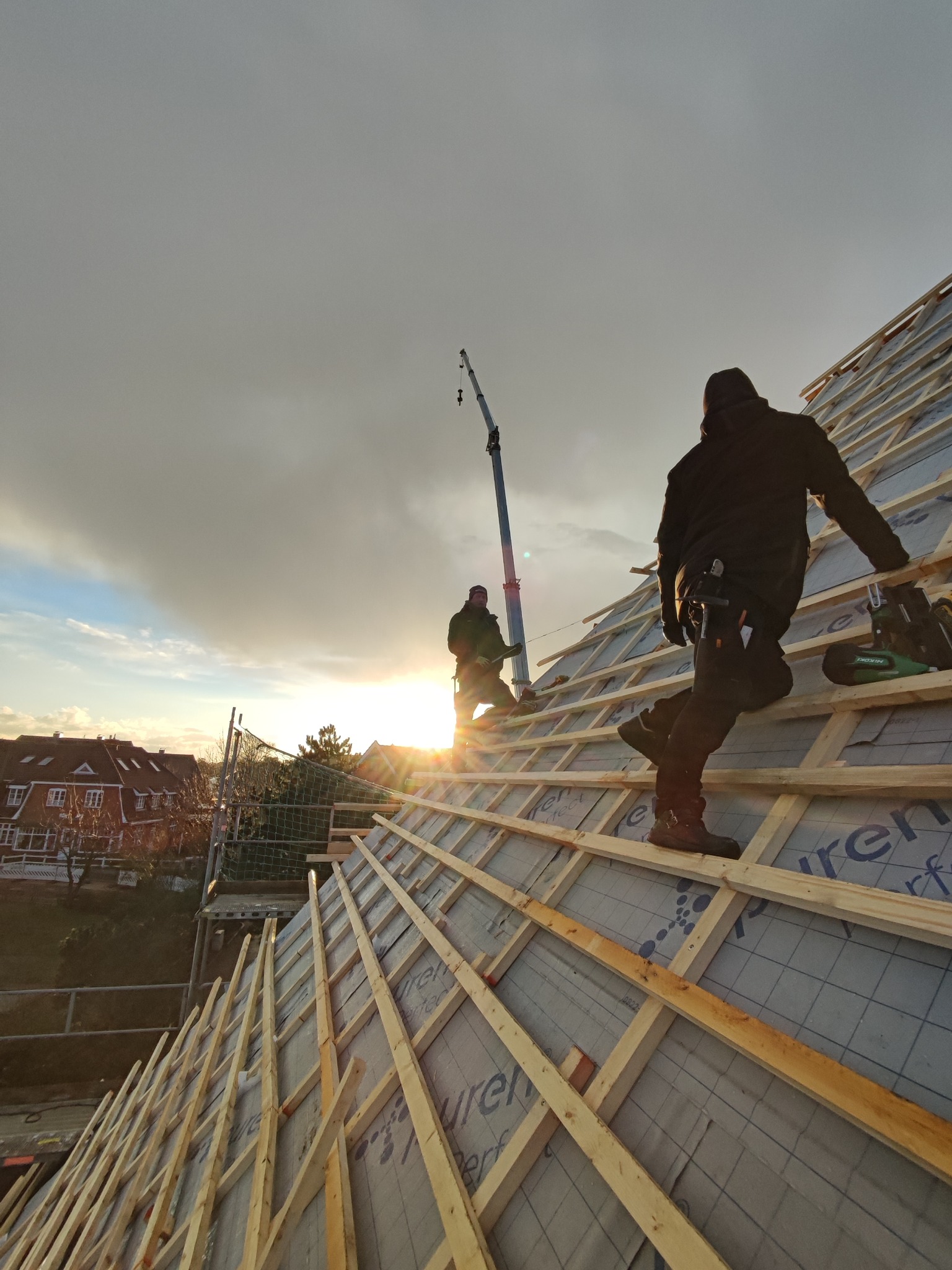 Dwóch dekarzy pracuje na drewnianej konstrukcji dachu w promieniach zachodzącego słońca, widoczny dźwig i zabudowa jednorodzinna w tle.