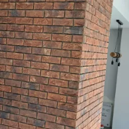 Dekoracyjny narożnik ściany wyłożony cegłą klinkierową w odcieniach czerwieni i brązu, widoczna lampa wisząca z elementami drewna i czerni, częściowo widoczny karton na podłodze.