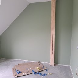 JW construction - Pokój na poddaszu z pomalowanymi na oliwkowo ścianami, surową drewnianą belką i fragmentami taśmy malarskiej na szarej podłodze.