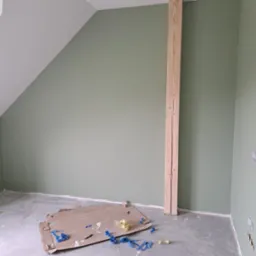 Pokój na poddaszu z pomalowanymi na oliwkowo ścianami, surową drewnianą belką i fragmentami taśmy malarskiej na szarej podłodze.