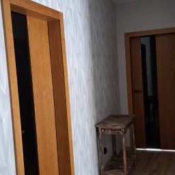 JW construction - Nowoczesny korytarz z drewnianymi drzwiami przesuwnymi, białą ścianą pokrytą geometrycznym wzorem, drewnianą podłogą i małym, postarzanym stolikiem z drewna.