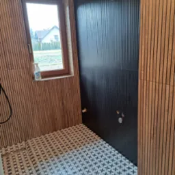 Nowoczesna łazienka w trakcie wykończenia: czarne płytki ścienne z pionowymi żłobieniami, drewniane panele ścienne, wzorzysta podłoga, okno z widokiem na dom i butelka płynu do czyszczenia...