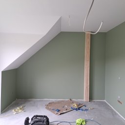 JW construction - Pokój po malowaniu ścian na oliwkowy kolor, widoczne niedoskonałości i pozostałości po remoncie, takie jak karton, taśma malarska i przewody elektryczne.