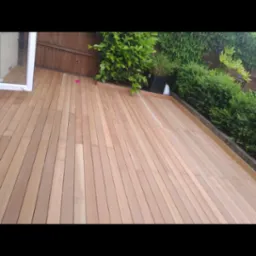 Nowo położony taras z desek kompozytowych w odcieniach ciepłego drewna, obrzeża wykończone, widok na zieleń ogrodu i fragment okna tarasowego.