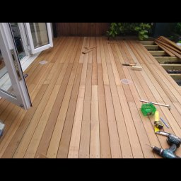 JW construction - Montaż drewnianego tarasu przy oknie balkonowym, widoczne narzędzia: wiertarka, miarka, poziomica, młotek, fragmenty desek i konstrukcja nośna.