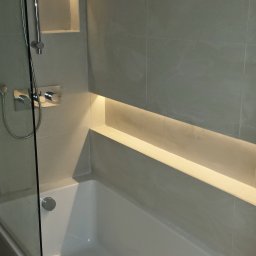 JW construction - Nowoczesna łazienka z wanną, szarą glazurą i podświetlaną wnęką w ścianie, widok na baterię prysznicową i szklany parawan.