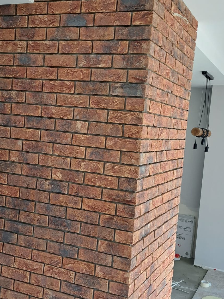 Dekoracyjny narożnik ściany wyłożony cegłą klinkierową w odcieniach czerwieni i brązu, widoczna lampa wisząca z elementami drewna i czerni, częściowo widoczny karton na podłodze.