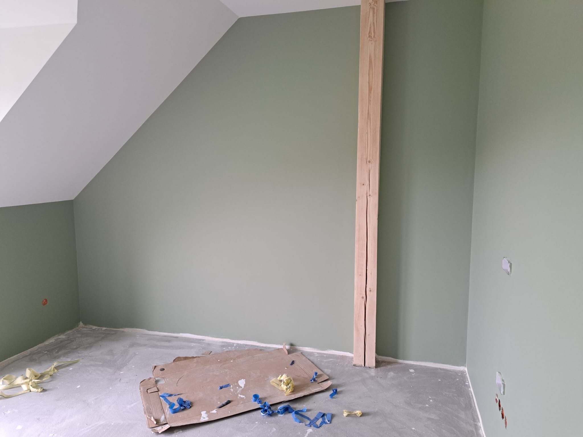 Pokój na poddaszu z pomalowanymi na oliwkowo ścianami, surową drewnianą belką i fragmentami taśmy malarskiej na szarej podłodze.