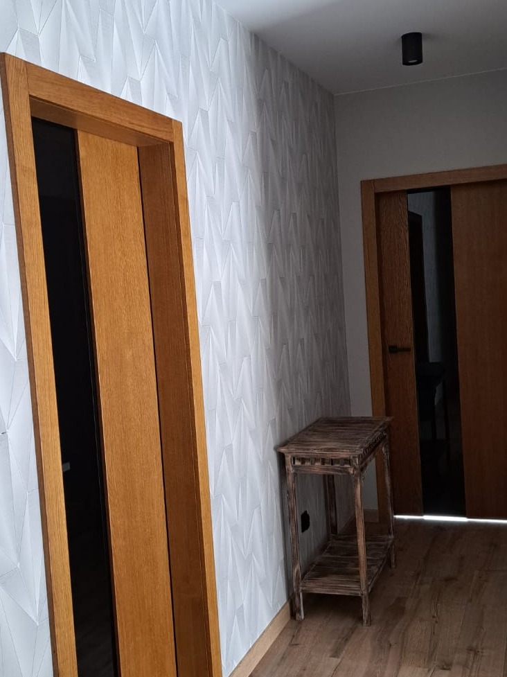 Nowoczesny korytarz z drewnianymi drzwiami przesuwnymi, białą ścianą pokrytą geometrycznym wzorem, drewnianą podłogą i małym, postarzanym stolikiem z drewna.