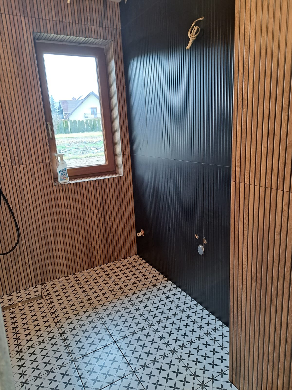 Nowoczesna łazienka w trakcie wykończenia: czarne płytki ścienne z pionowymi żłobieniami, drewniane panele ścienne, wzorzysta podłoga, okno z widokiem na dom i butelka płynu do czyszczenia...