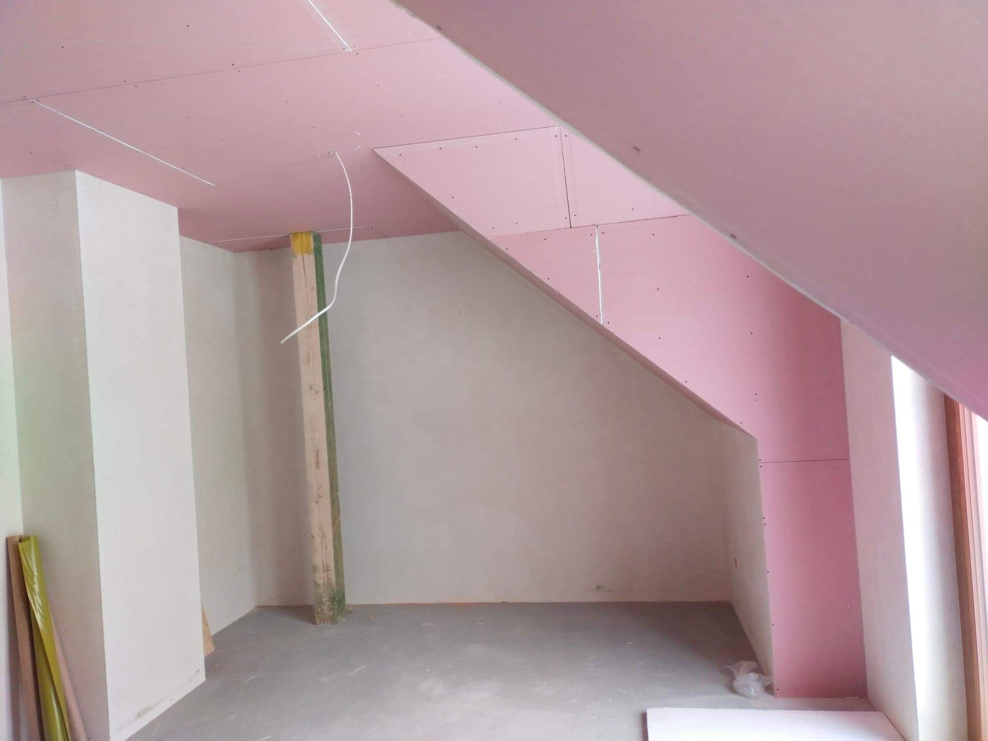 Montaż płyt gipsowo-kartonowych na poddaszu, widoczne profile i przewody elektryczne, surowy beton na podłodze, fragment okna w prawym dolnym rogu.