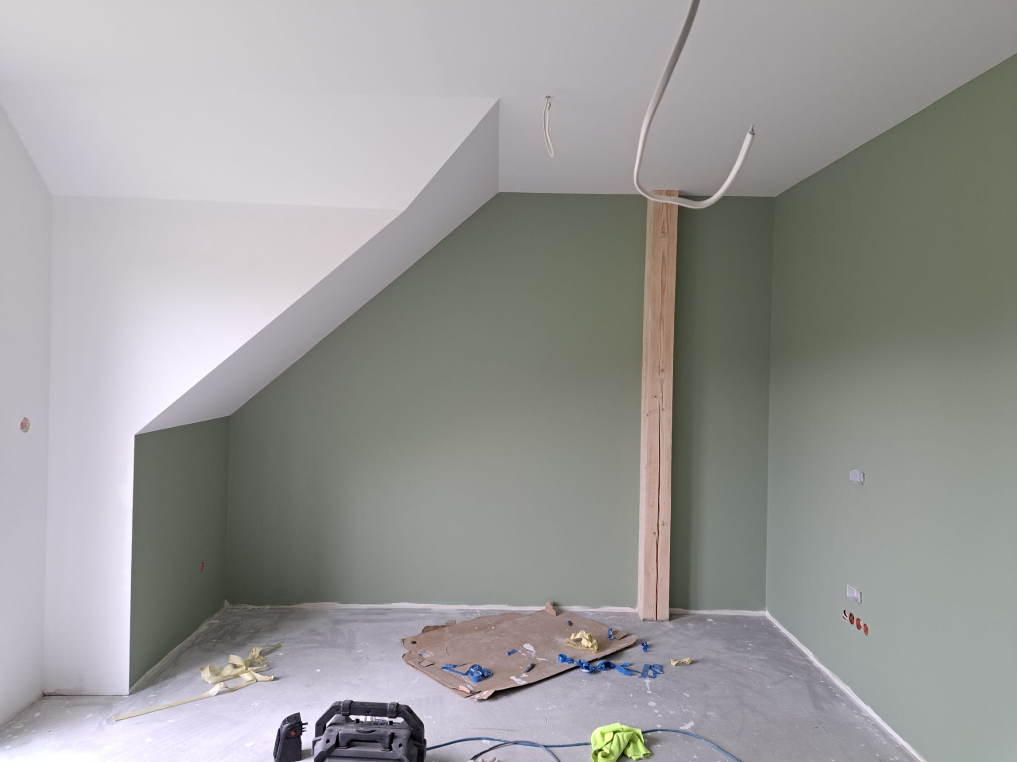Pokój po malowaniu ścian na oliwkowy kolor, widoczne niedoskonałości i pozostałości po remoncie, takie jak karton, taśma malarska i przewody elektryczne.