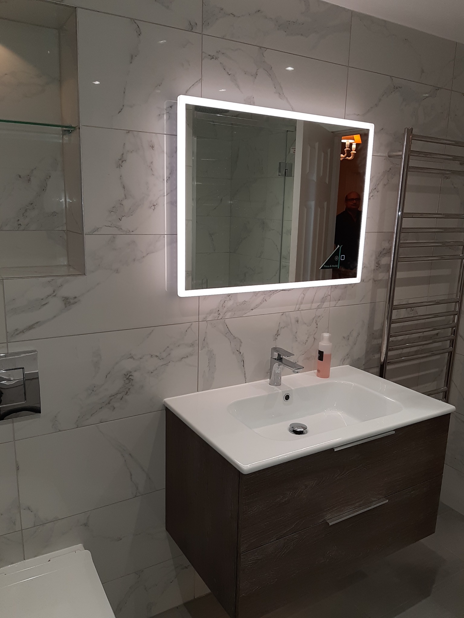 Nowoczesna łazienka z umywalką na szafce z ciemnego drewna, podświetlanym lustrem i marmurowymi płytkami na ścianach, widoczny grzejnik drabinkowy i fragment toalety.