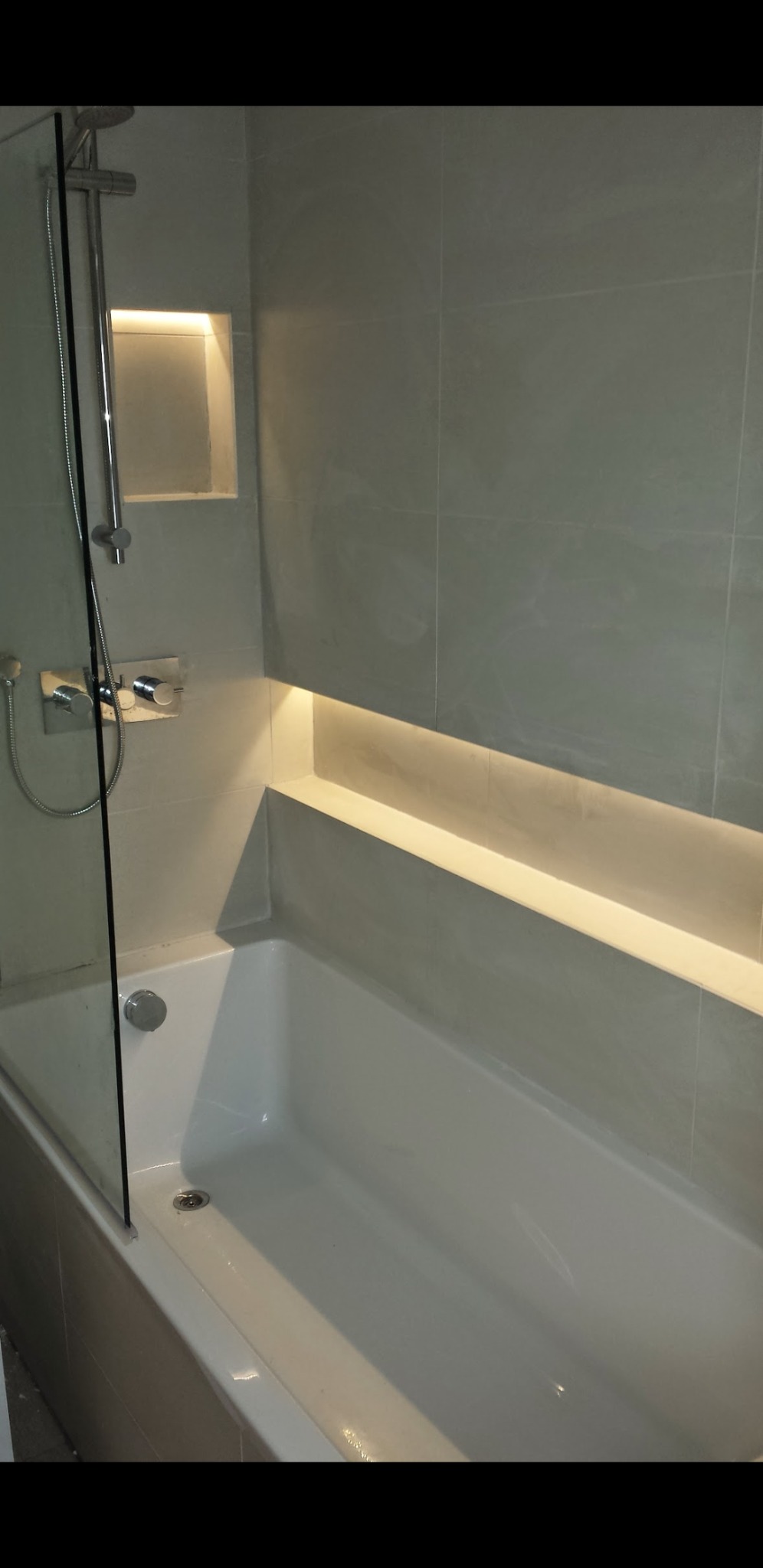 Nowoczesna łazienka z wanną, szklanym parawanem nawannowym i podświetlanymi wnękami w szarych płytkach.