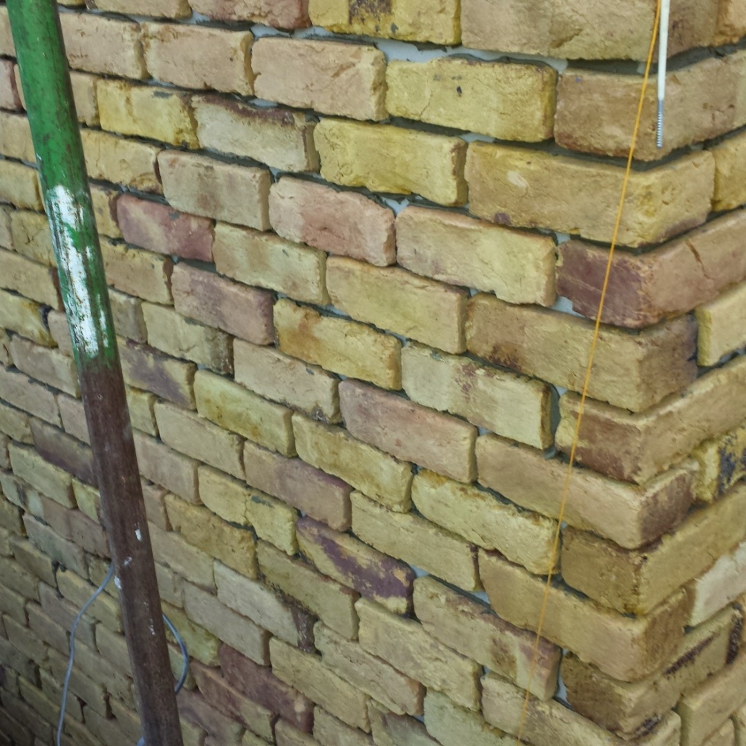 Żółto-czerwona ściana z cegły klinkierowej z zieloną rurą i żółtą nicią pionu murarskiego w rogu.