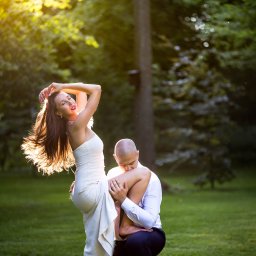 Paweł Tawkin fotografia ślubna - Romantyczna sesja w plenerze: mężczyzna klęczy, całując nogę kobiety w białej sukni na tle zieleni i promieni słonecznych. Dynamiczne ujęcie, podkreślone światłem.
