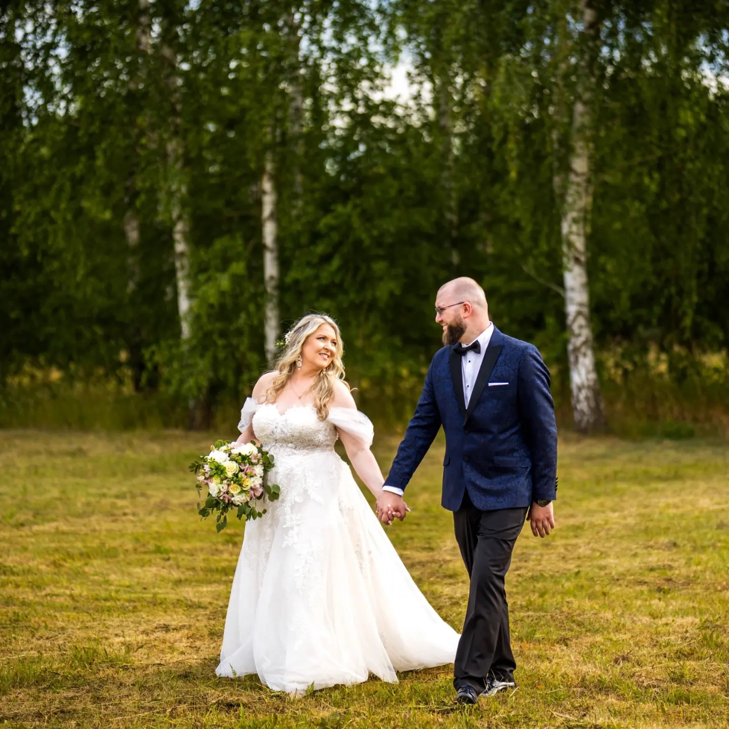 Para młoda, on w granatowej marynarce z delikatnym wzorem, ona w białej sukni ślubnej z bukietem polnych kwiatów, spacerują po łące z brzozowym zagajnikiem w tle.