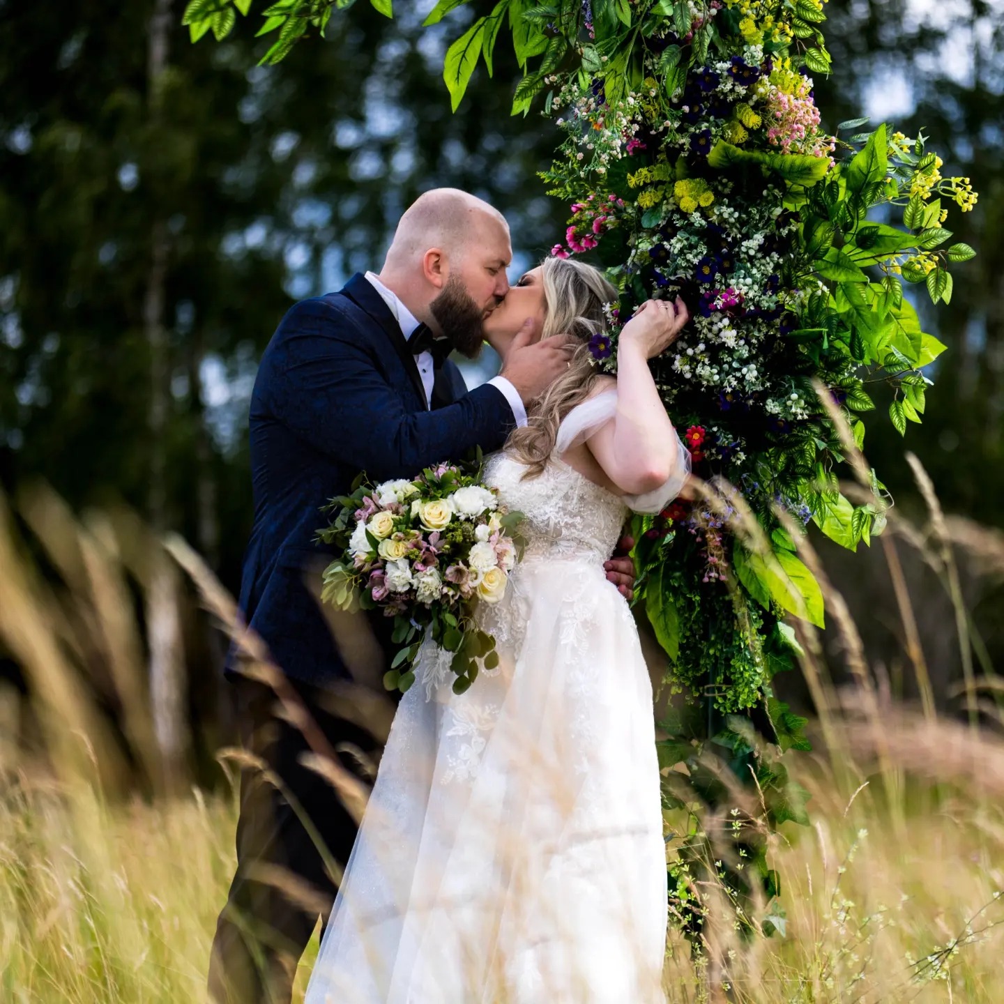 Para młoda całuje się pod łukiem kwiatowym w plenerze, panna młoda w koronkowej sukni trzyma bukiet, a pan młody w granatowym garniturze w delikatny wzór.