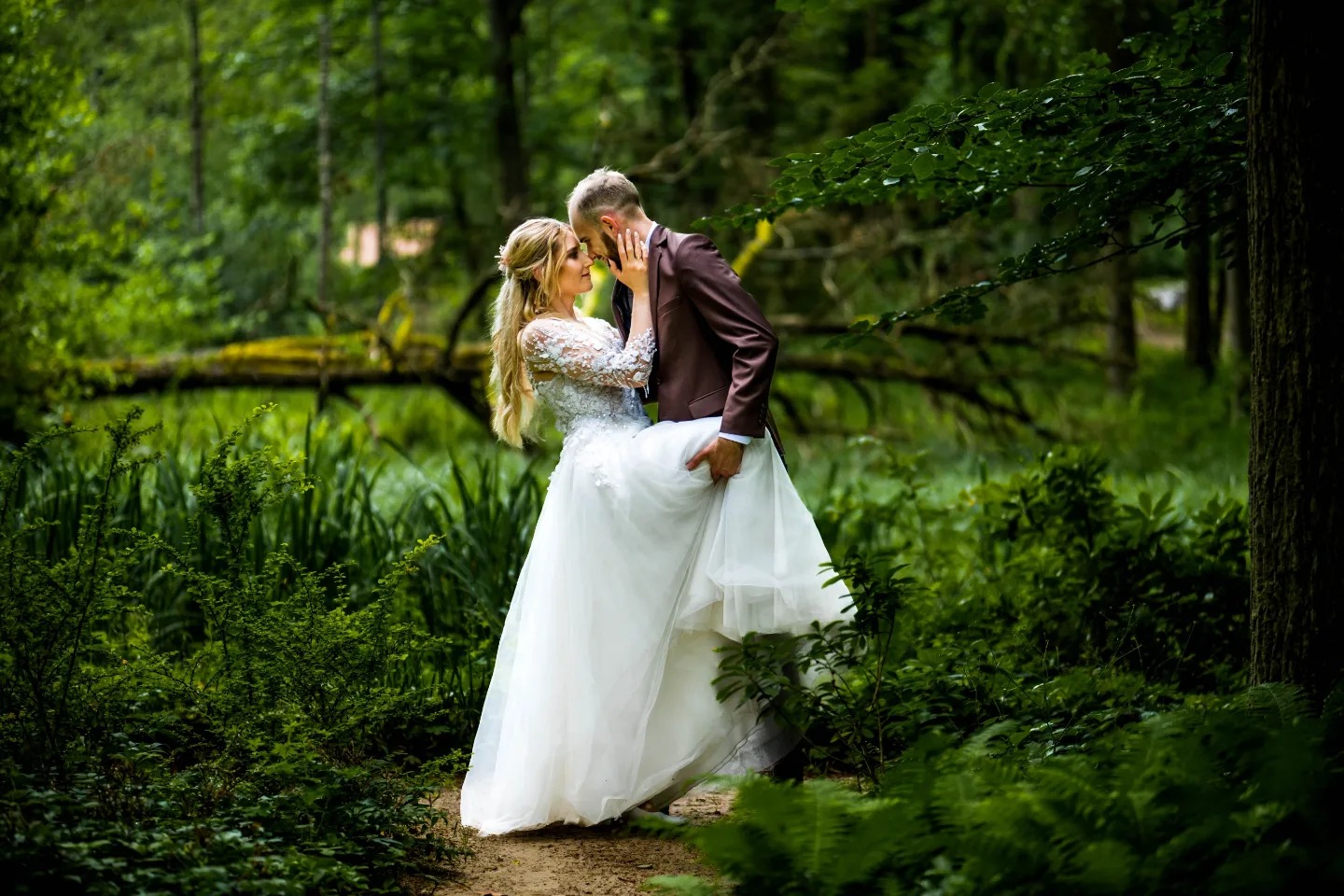 Para młoda w lesie: Panna młoda w koronkowej sukni z długim rękawem, podtrzymywana przez pana młodego w brązowym garniturze, oboje patrzą sobie w oczy na tle zieleni.