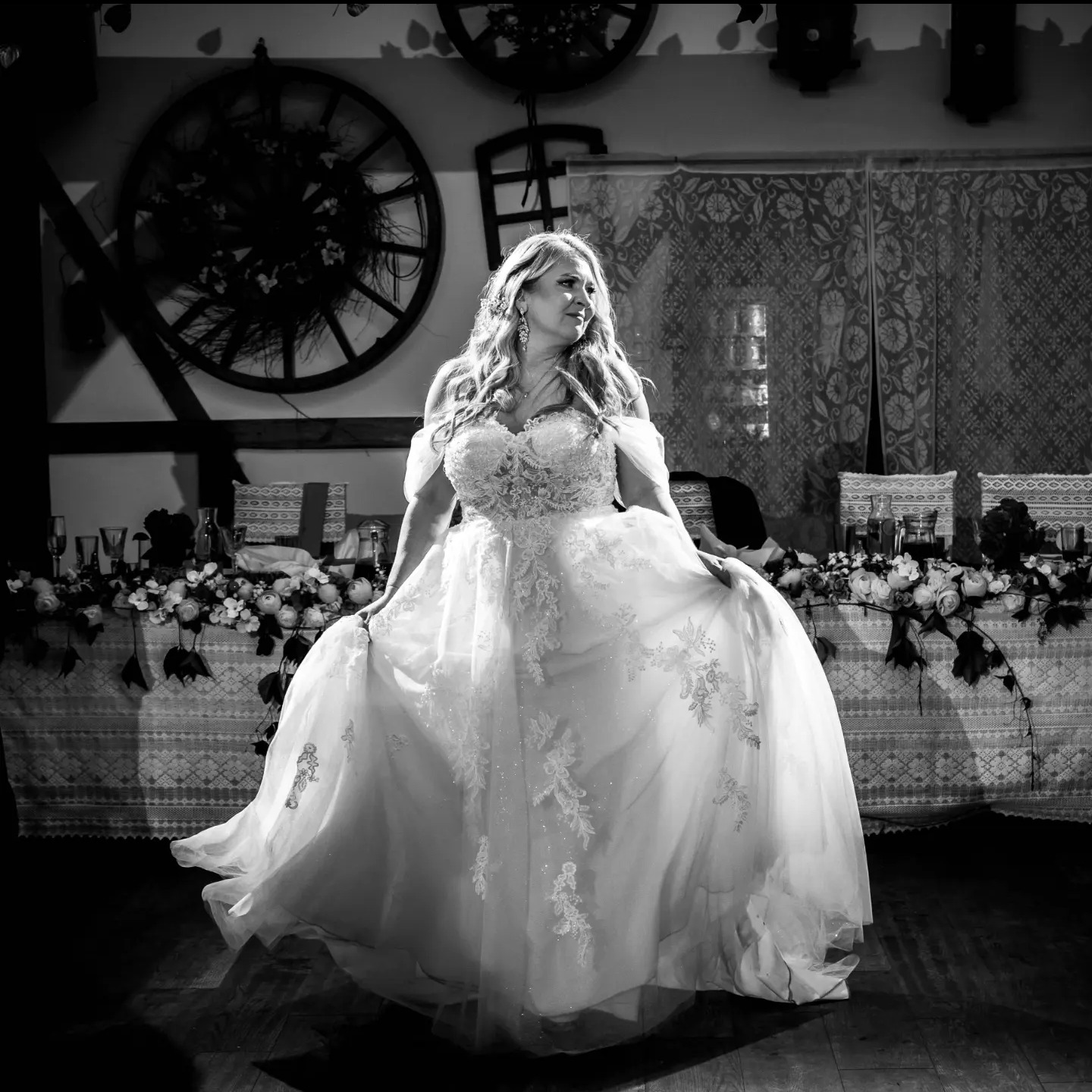 Panna młoda w koronkowej sukni, pozująca na tle dekoracji weselnych z drewnianymi kołami i kwiatami, czarno-białe zdjęcie.