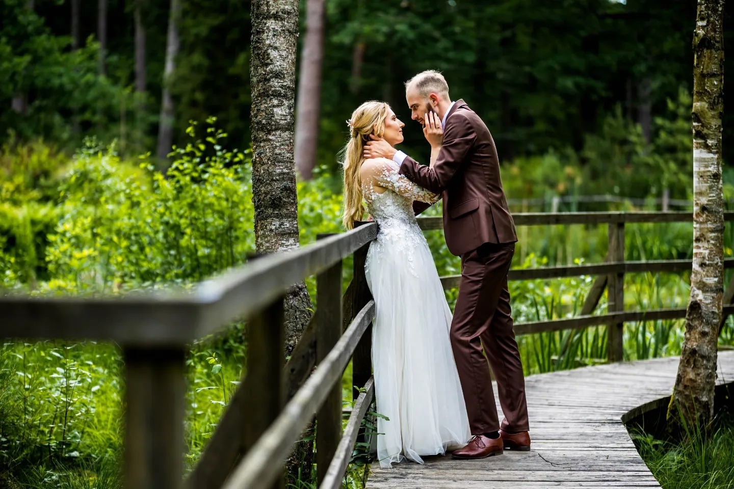 Para młoda: blondwłosa kobieta w koronkowej sukni ślubnej i mężczyzna w brązowym garniturze, stojący na drewnianej kładce w otoczeniu zieleni, w czułym uścisku.