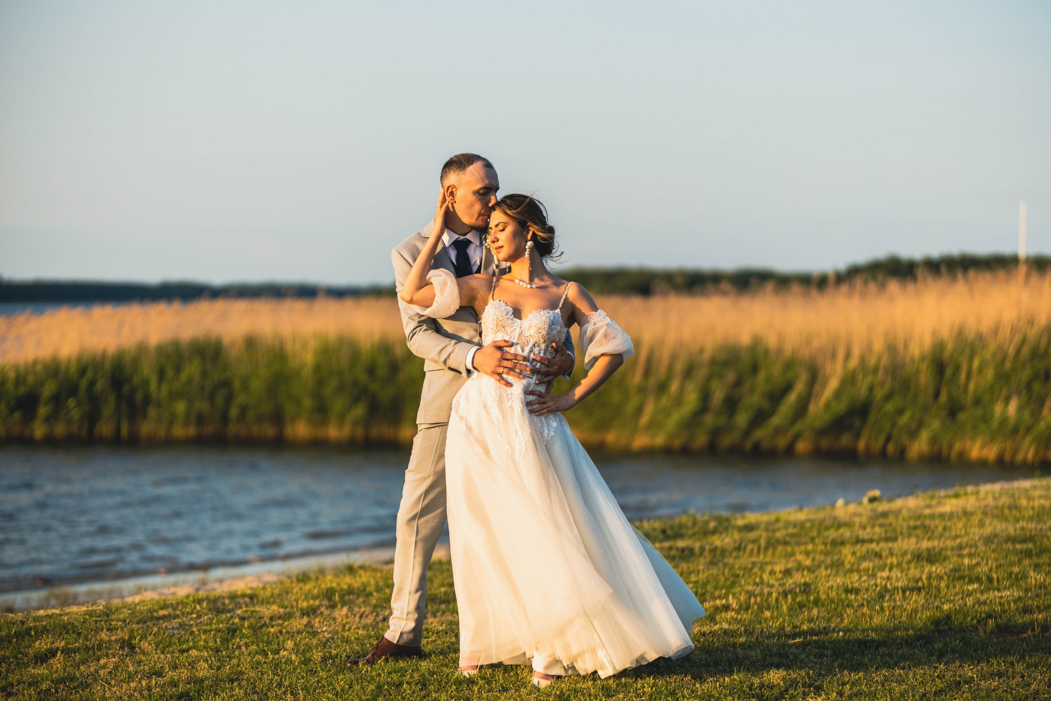 Para młoda w objęciach na tle jeziora i trzcin. Jasna suknia z koronką, szary garnitur. Romantyczny plener, ciepłe światło zachodzącego słońca.
