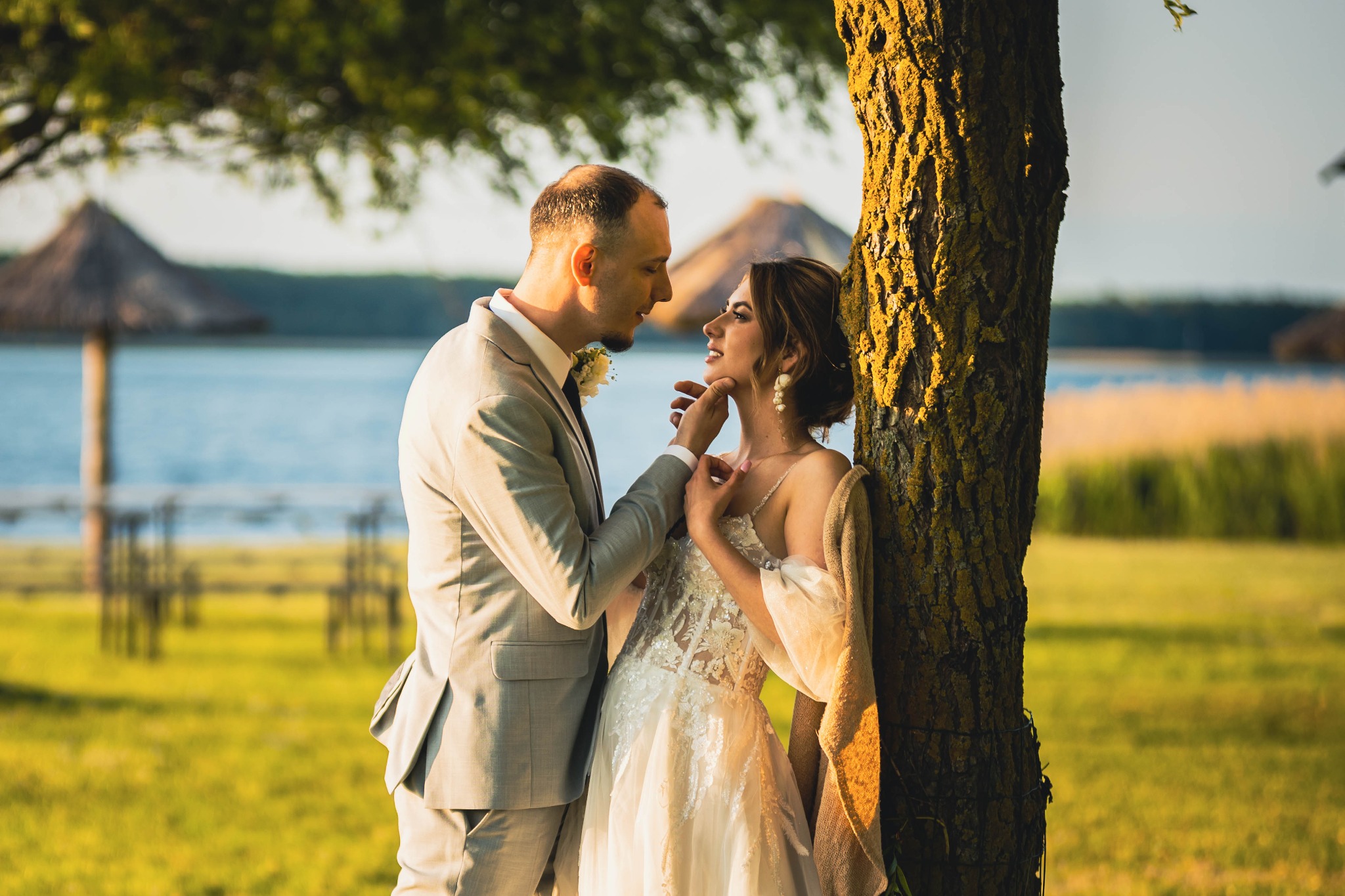 Para młoda oparta o drzewo, jezioro w tle. Pan młody w szarym garniturze dotyka twarzy panny młodej w białej sukni. Romantyczna sesja plenerowa.