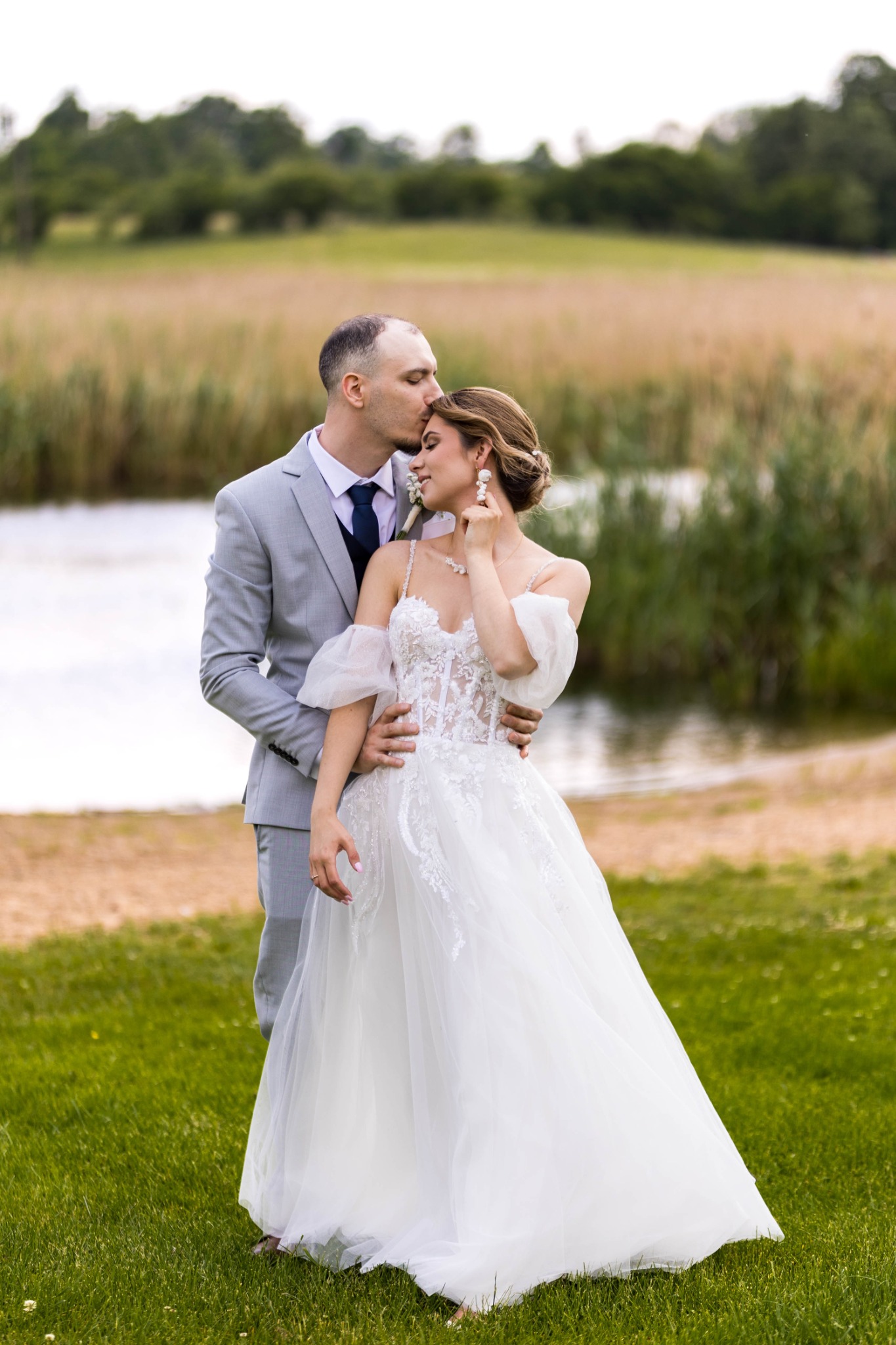 Para młoda w plenerze: mężczyzna w szarym garniturze całuje kobietę w białej sukni w czoło. W tle jezioro i łąka. Romantyczna sesja ślubna.