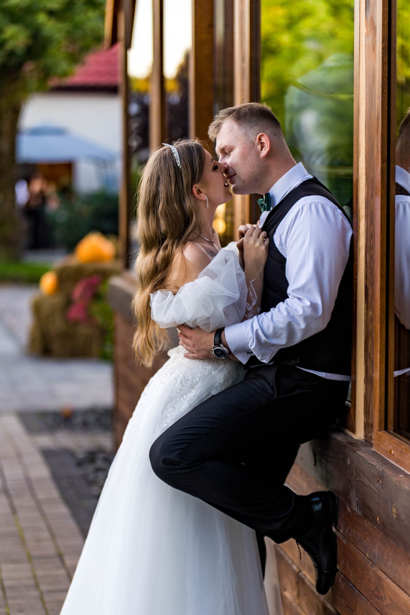 Pocałunek nowożeńców opartych o drewnianą konstrukcję z oknami. Panna młoda w białej sukni, pan młody w czarnym garniturze i zielonej muszce. W tle dekoracje ślubne.