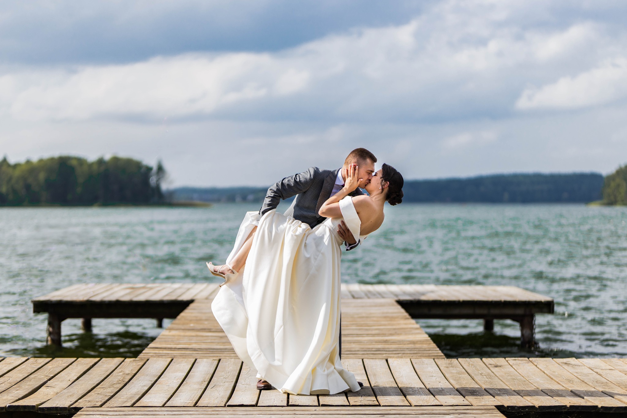 Para młoda na drewnianym pomoście nad jeziorem. Pan młody trzyma pannę młodą w ramionach, całując ją. W tle woda i zalesiony brzeg pod pochmurnym niebem.